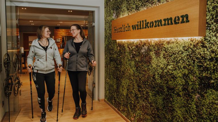Zwei Frauen gehen mit Nordic-Walking-Stöcken in einen freundlich gestalteten Empfangsbereich. Im Hintergrund hängt ein Willkommensschild an einer begrünten Wand.