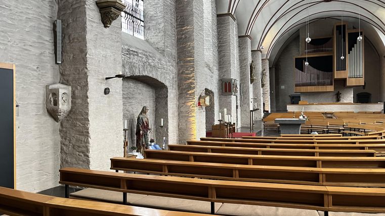 A quiet church with wooden benches and a high ceiling. On the wall, there are religious statues and windows visible.