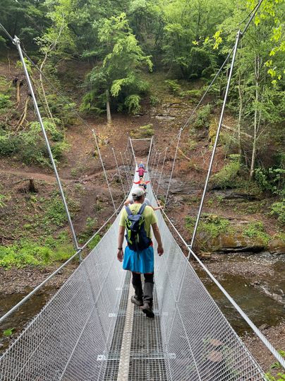 Een hangbrug over een kleine beek in een groene bos. Een persoon loopt langzaam over de brug.