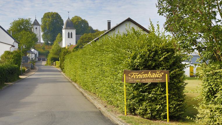 Ein ruhiger Weg mit grünen Hecken und einem Ferienhaus-Schild. Im Hintergrund sind zwei Kirchen zu sehen, umgeben von Bäumen.