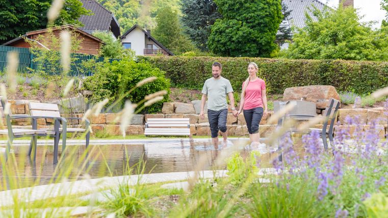 Ein Paar spaziert entspannt entlang eines Gartenteichs. Umgeben von grünen Pflanzen und Bänken bietet die Anlage eine ruhige und natürliche Atmosphäre.