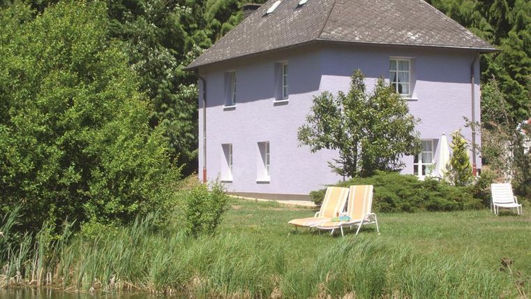 A cozy purple house surrounded by trees and greenery. In the foreground is a small pond with loungers for relaxing.