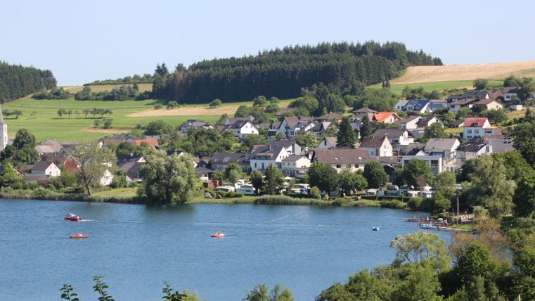 Ein ruhiger See umgeben von grünen Wäldern und einer kleinen Siedlung. Einige Boote sind auf dem Wasser sichtbar.