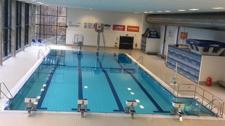 Een modern zwembad met helder water en startblokken. Veel daglicht valt door grote ramen de ruimte binnen.
