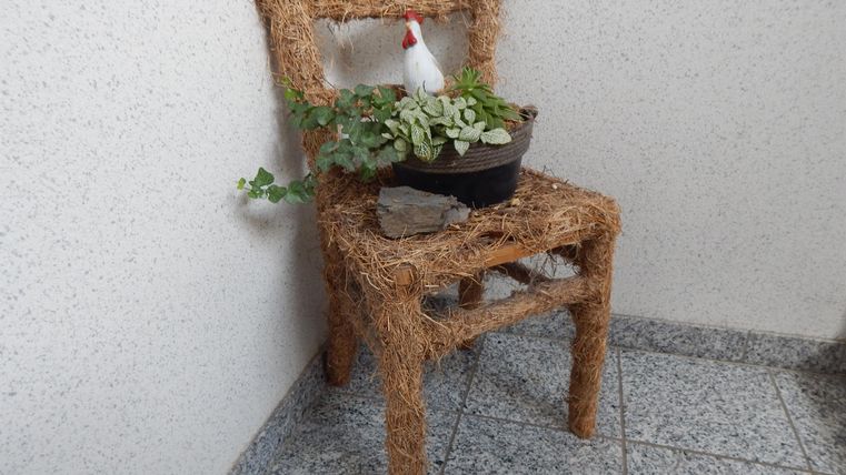 A chair made of natural materials stands in a corner. On the chair, there is a pot with plants and a small figure.