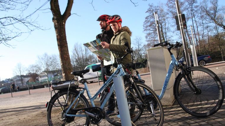 A pair of cyclists is standing next to their bicycles and looking at a map. The surroundings are bright, with trees and a clear view.