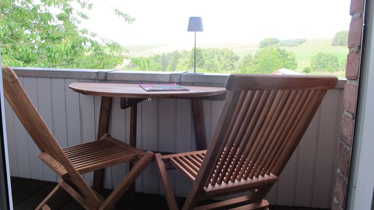 Ein gemütlicher Balkon mit einem kleinen Tisch und zwei Holzstühlen. Im Hintergrund sind grüne Bäume und eine weite Landschaft zu sehen.