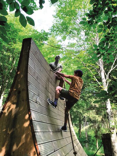 Een jonge man klimt aan een houten wand in een bos. Op de achtergrond zijn groene bomen te zien.