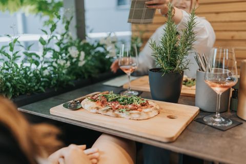 Ein köstliches Fladenbrot mit frischem Gemüse und Kräutern auf einem Holzbrett. Dahinter stehen Gläser mit Rosé-Wein und eine grüne Pflanze auf dem Tisch.