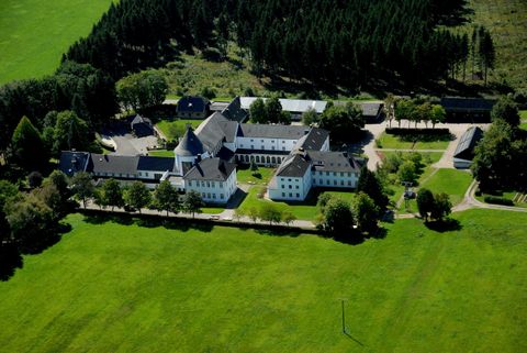 Eine große weiße Gebäudeanlage liegt von grünen Wiesen umgeben. Im Hintergrund sind Bäume und ein Wald sichtbar.