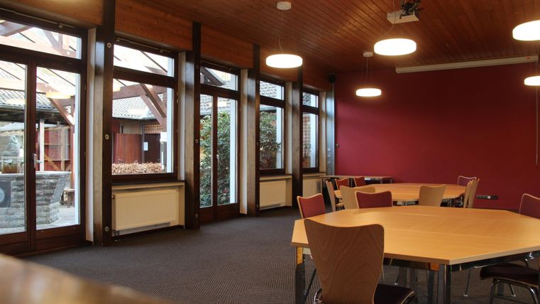 A bright room with large windows and a wooden ceiling. In the room, there are several tables and chairs.