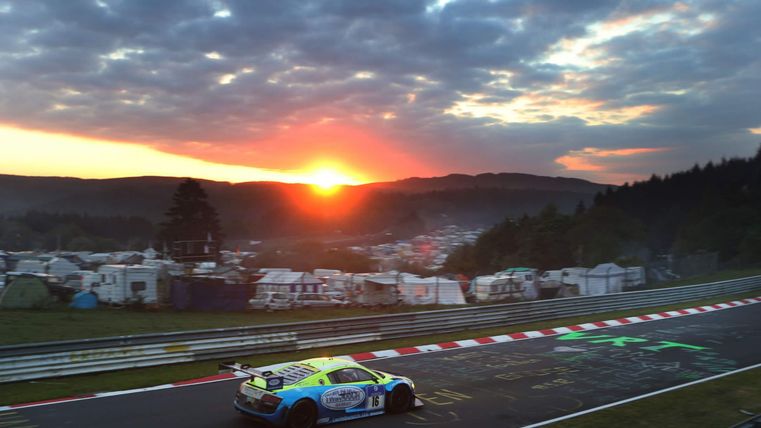 Een raceauto rijdt bij zonsondergang op een bekende racebaan. Op de achtergrond zijn tentplaatsen en een pittoresk landschap te zien.