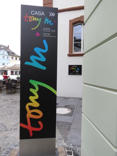 Ein modernes Schilderdesign mit bunten Schriftzügen zeigt den Namen "Casa Tony M". Das Schild steht an einer belebten Straßenecke.