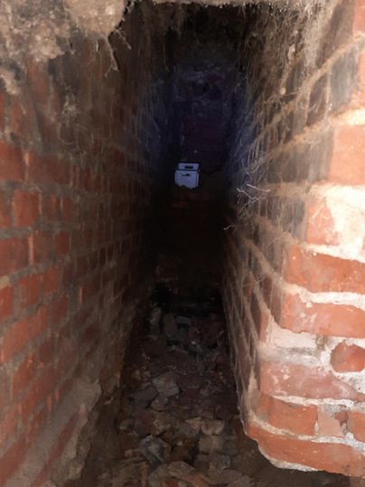 A narrow, dark-looking tunnel made of bricks with stones and debris on the floor. At the end of the tunnel, a faint light is visible.