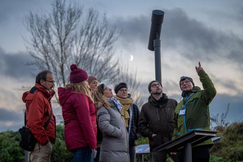Een groep mensen staat buiten en luistert naar een spreker die naar een punt wijst. De lucht is bewolkt en het is schemering.