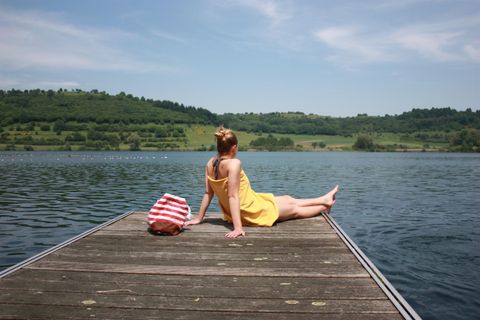 Eine Frau in einem gelben Kleid sitzt entspannt auf einem Holzsteg am Wasser. Im Hintergrund sind grüne Hügel und ein klarer Himmel zu sehen.