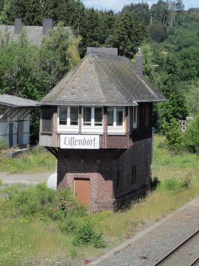 View from above of an old building about 3 meters wide.
The narrow ground floor consists of red bricks.
On top of it is a wooden structure made of brown boards, which is slightly wider than the ground floor.
In the lower area, there is a brown door.
The upper area has three white windows at the front.
Below the windows, there is a white sign with the inscription Lissendorf.
The roof of the signal box is covered with black slate.