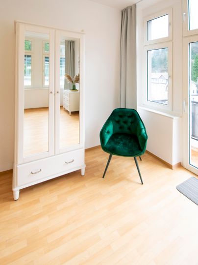 Ein helles Zimmer mit einem großen Spiegelkasten und einem grünen Sessel. Die Fenster lassen viel natürliches Licht herein.