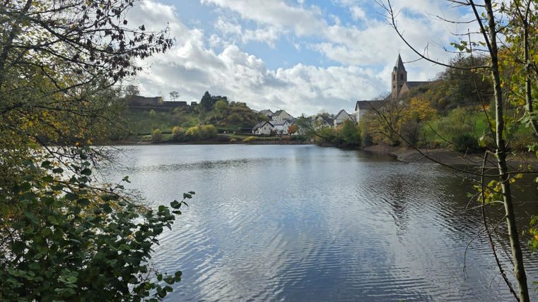 Een rustige zee omgeven door bomen en kleurrijk herfstblad. Op de achtergrond zijn huizen en een kerk zichtbaar.
