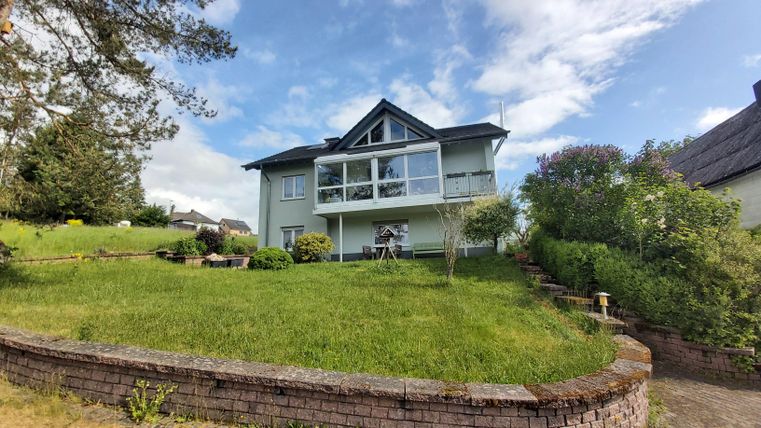 Een mooi huis met een grote tuin en veel groen. De lucht is helder en er zijn enkele bomen in de omgeving.