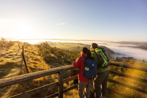 Ein Paar steht an einer Aussichtplattform und blickt auf eine nebelige Landschaft im Morgenlicht. Die Szenerie zeigt sanfte Hügel und einen klaren Himmel.