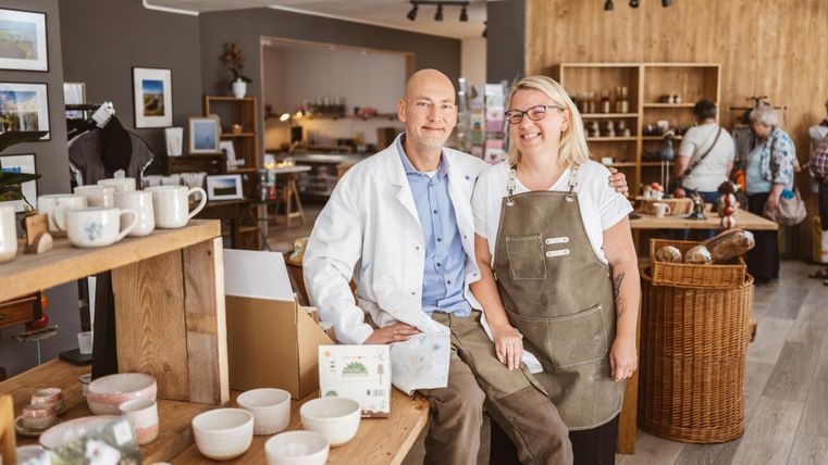 A man in a white coat and a woman in an apron are smiling next to each other in a store. The store is filled with ceramics and other goods.