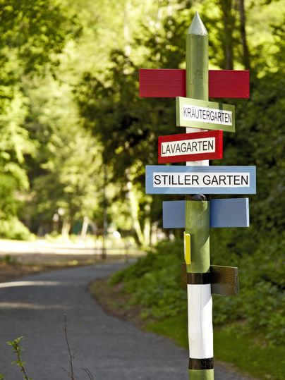 Ein Wegweiser zeigt die Richtungen zu verschiedenen Gärten: Kräutergarten, Lavagarten und Stiller Garten. Die Umgebung ist grün und einladend.