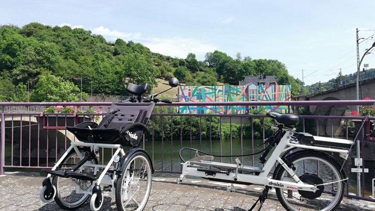 Ein adaptives Fahrrad steht auf einer Brücke mit Blick auf einen Fluss. Im Hintergrund sind Bäume und eine farbenfrohe Wandmalerei zu sehen.