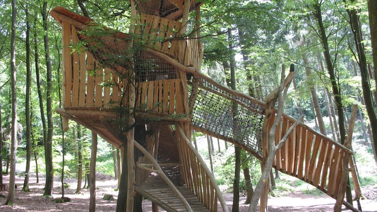 Ein Spielbaumhaus aus Holz steht in einem Waldgebiet. Es hat mehrere Etagen und eine Rutsche, um Spaß zu bieten.
