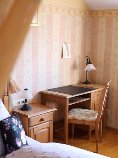 Ein gemütliches Zimmer mit einem schlichten Holzschreibtisch, einem Stuhl und einer Lampe. Die Wände sind mit zartem, gestreiftem Papier in Pastellfarben dekoriert.