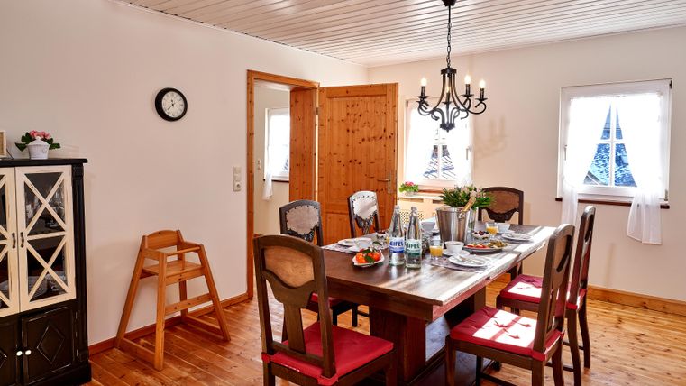 An inviting dining area with a large wooden table and red chairs. The room has bright walls and large window light.