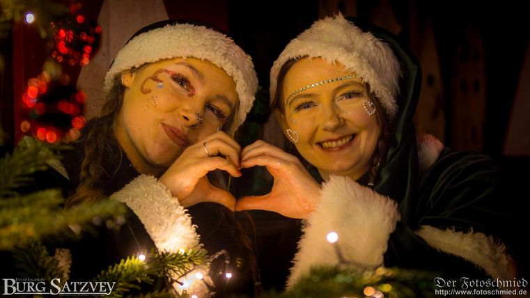 Zwei fröhliche Frauen in Weihnachtskleidung formen mit ihren Händen ein Herz. Im Hintergrund sind festliche Lichter und Tannenzweige zu sehen.