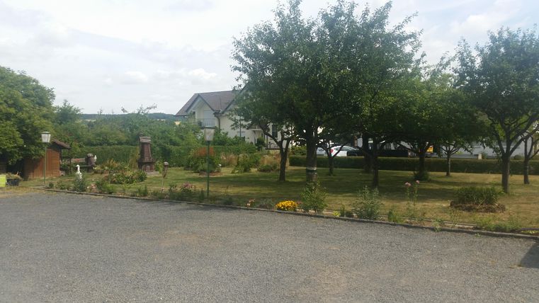 Ein ruhiger Garten mit großen Bäumen und bunten Blumen. Im Hintergrund sieht man ein kleines Haus und eine weitläufige Landschaft.
