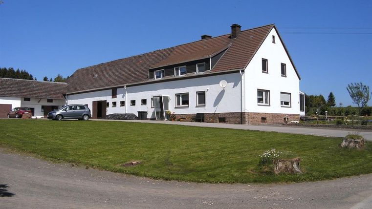 Ein großes, weißes Haus mit einem zentralen Eingang und einem roten Dach. Der Garten ist gepflegt und die Umgebung ist grün und ländlich.