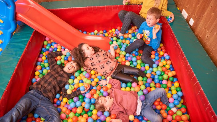 Eine fröhliche Gruppe von Kindern liegt in einem Ballbecken mit bunten Bällen. Im Hintergrund ist eine Rutsche zu sehen.
