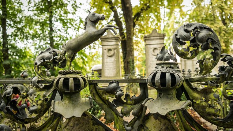 Een kunstzinnig smeedijzeren poort met een springende hondfiguur. Op de achtergrond zijn monumentale grafstenen en groene bomen zichtbaar.