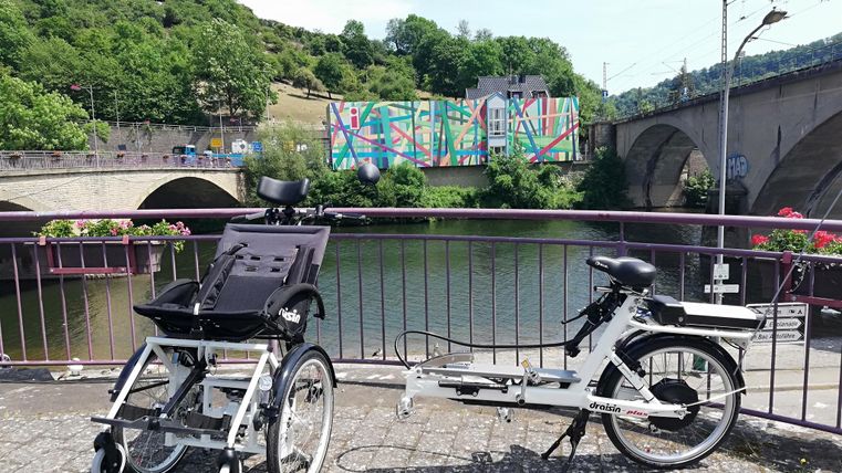 Ein Rollstuhl und ein Fahrrad stehen an einem Flussufer. Im Hintergrund sind bunte Wandmalereien und eine Brücke zu sehen.