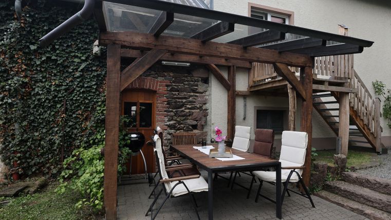 Ein gemütlicher Außenbereich mit einem Holzpergola, einem Tisch und Stühlen. Im Hintergrund ist ein Gebäude mit einer Treppe zu sehen.