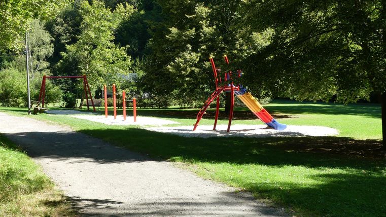 Ein Spielplatz mit einer Rutsche, Schaukeln und mehreren Spielgeräten steht auf einem grünen Rasen. Der Bereich ist von Bäumen umgeben und gibt einen einladenden Eindruck.