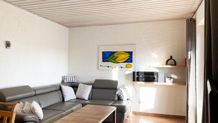 Ein modernes Wohnzimmer mit einem grauen Sofa und einem Holztisch. An der Wand hängt ein farbenfrohes Bild, und es gibt viel natürliches Licht.