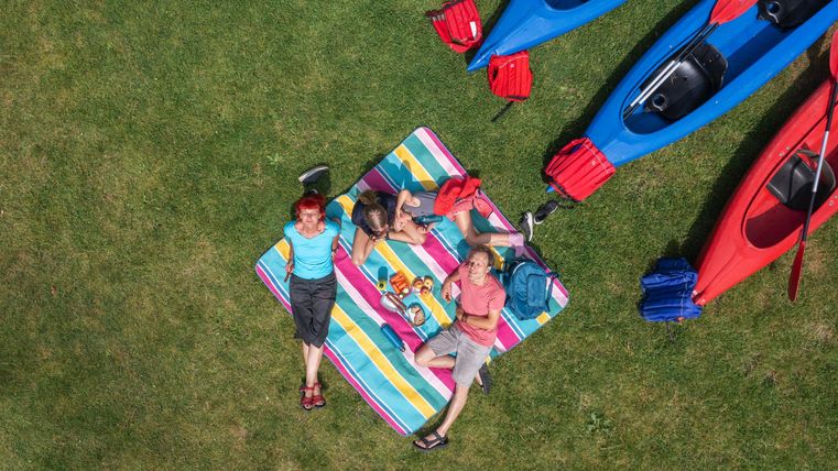 Drei Personen liegen auf einer bunten Decke auf dem Gras. Neben ihnen stehen mehrere Kajaks in Blau und Rot.