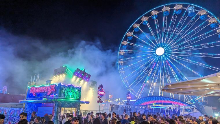 Ein festlicher Jahrmarkt mit vielen Menschen und bunten Lichtinstallationen. Im Hintergrund steht ein großes Riesenrad, das in der Nacht leuchtet.