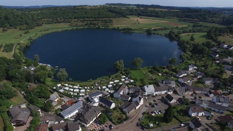 Ein ruhiger See umgeben von einer malerischen Landschaft und einem kleinen Dorf. Die Gegend ist grün und einladend, mit klaren Himmel und weitläufigen Feldern.