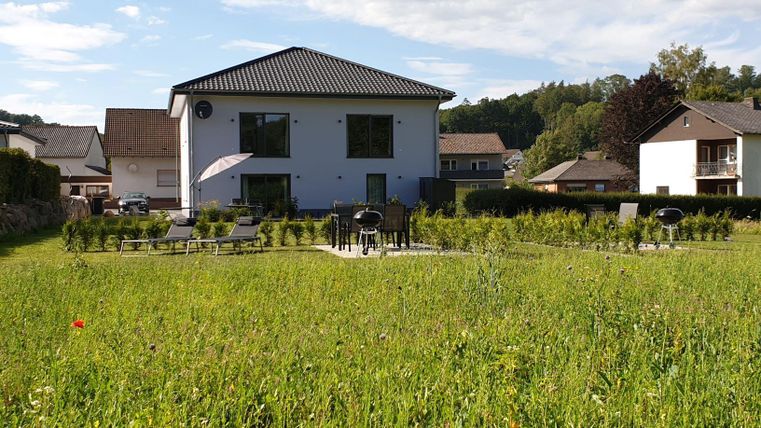 Ein modernes Haus umgeben von einer grünen Wiese. Im Vordergrund sind Gartenmöbel und eine einladende Terrasse zu sehen.