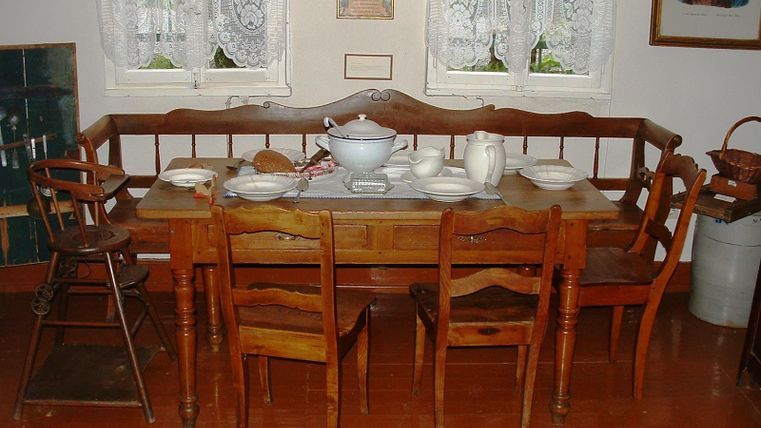 Ein nostalgischer Essbereich mit einem großen Holztisch und sechs Stühlen. Auf dem Tisch stehen Geschirr und eine Suppenplatte, umgeben von hellen Fenstern mit Vorhängen.