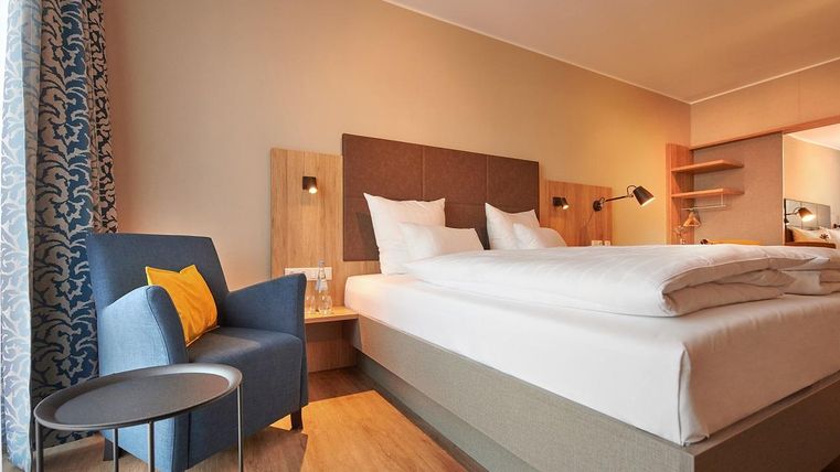 Ein modernes Hotelzimmer mit einem Doppelbett und einem blauen Sessel. Das Zimmer hat helle Farben und eine einladende Atmosphäre.