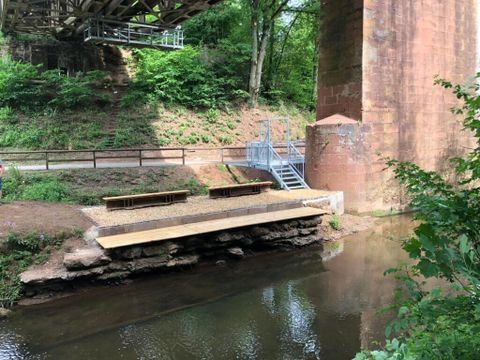 Ein ruhiger Wasserbereich unter einer Brücke, umgeben von grünem Bewuchs. Eine kleine Holzbank lädt zum Verweilen ein.