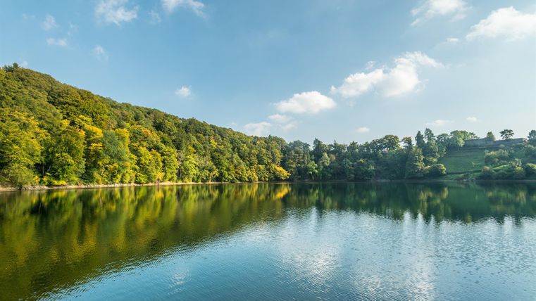 Ein ruhiger See umgeben von grünen Bäumen und sanften Hügeln. Der Himmel ist klar mit einigen Wolken.