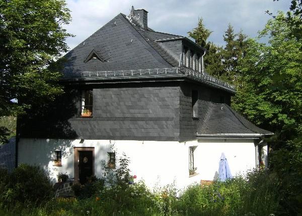 Een charmant huis met zwarte shingles en een witte onderbouw. Het is omringd door bomen en bloeiende planten.