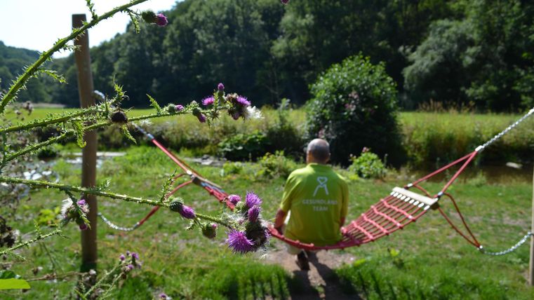 Eine Person sitzt auf einer Hängematte in der Natur. Im Hintergrund sind Bäume und ein ruhiger Fluss zu sehen.